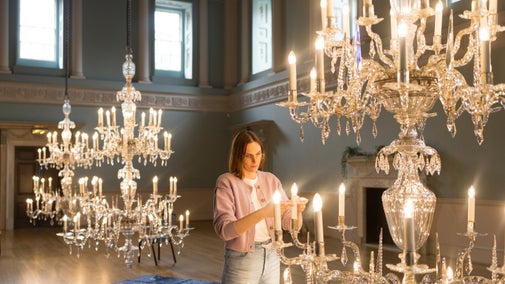 Conservator stand in a large room with high windows, looking closely at the glass on a lit chandelier that has been brought down from the ceiling.